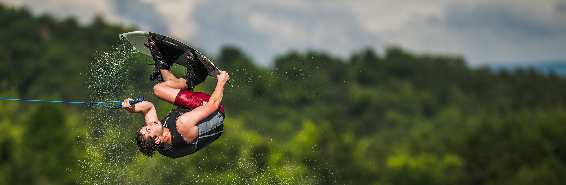 Person wakeboarding and doing a flip against a background of trees and cloudy sky.