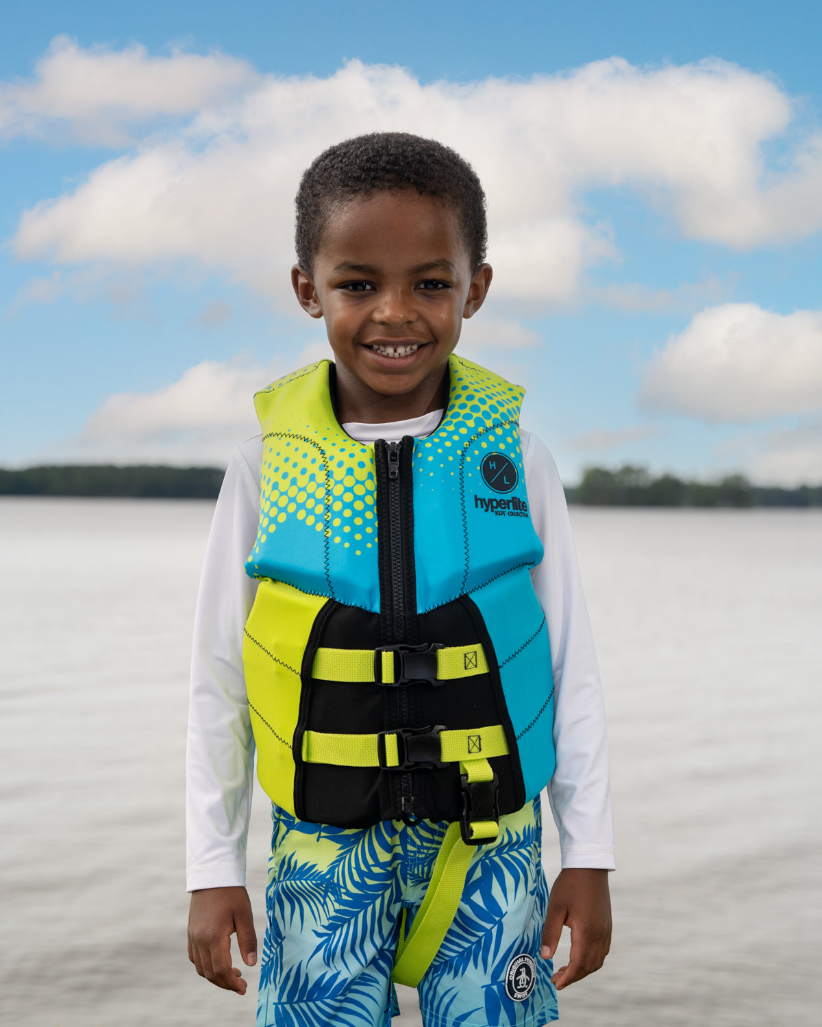 Smiling child in a colorful life jacket standing by a lake.