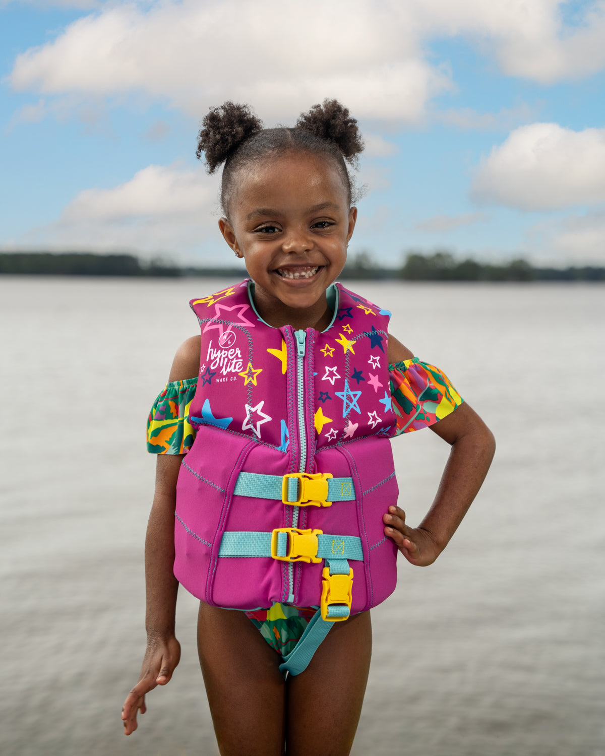 Child smiling in colorful life jacket by a lake.