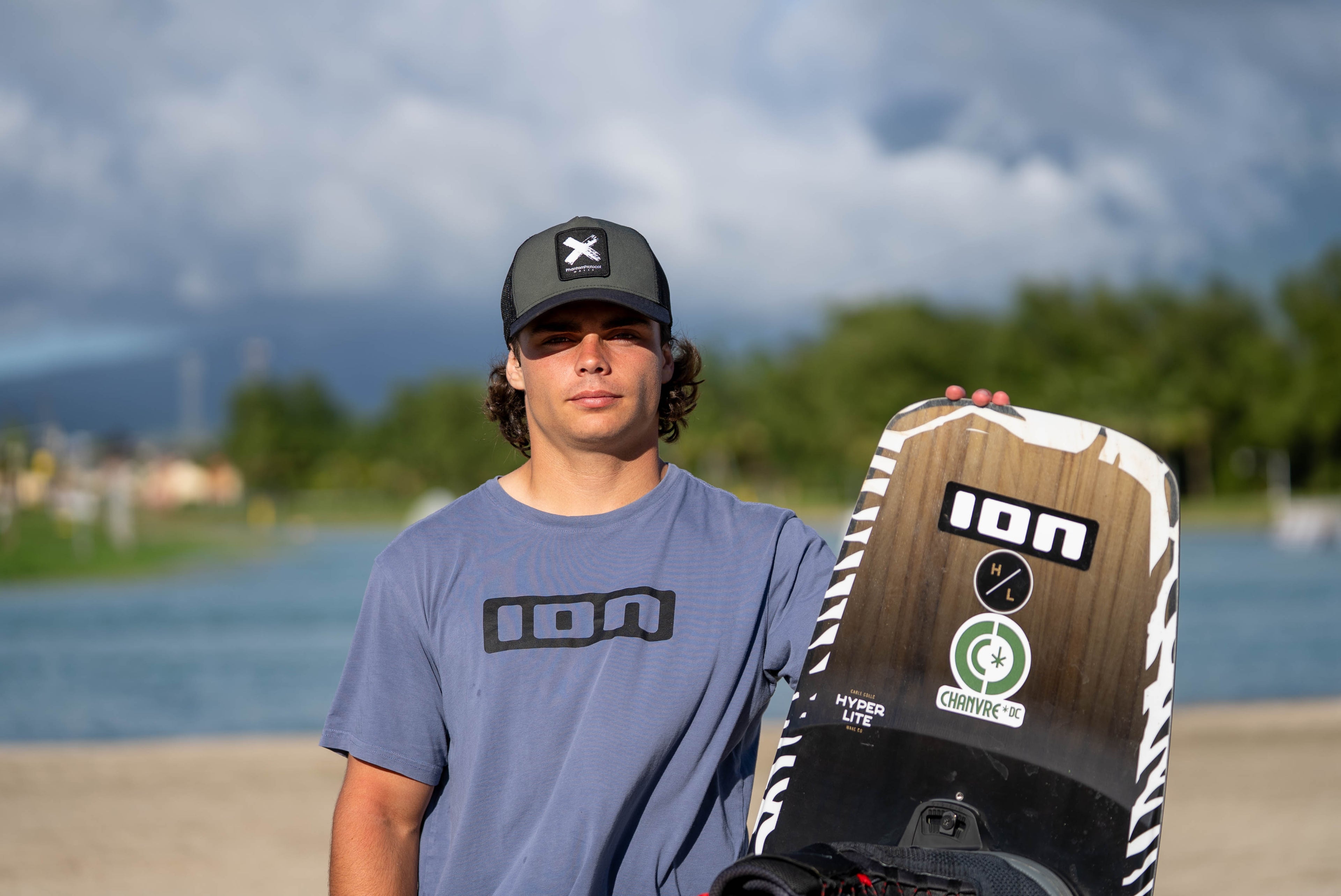 Person standing outdoors with a wakeboard near a body of water, wearing a cap and t-shirt.