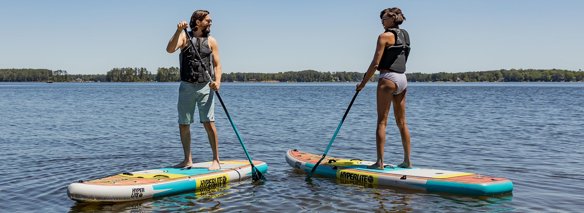 Two people paddleboarding on a lake under a clear sky, wearing life jackets.