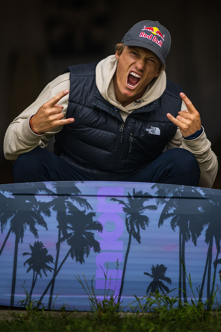 Person in a cap doing rock hand signs, standing behind a surfboard with palm tree design.