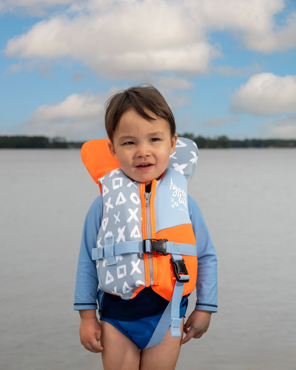 Young child in a life jacket stands near a calm lake under a cloudy sky.