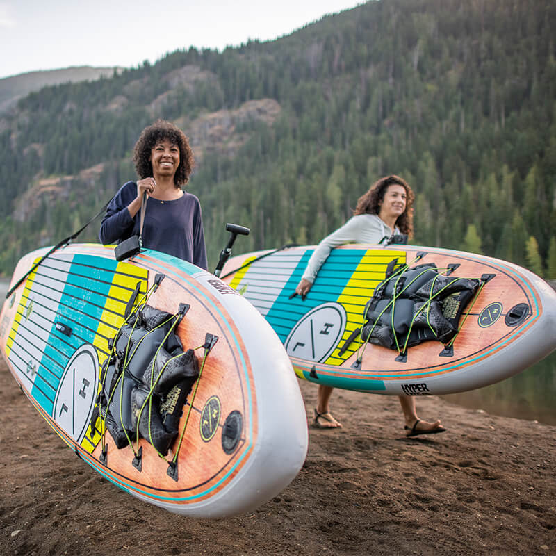 Two people carrying paddleboards by a scenic, forested lake.