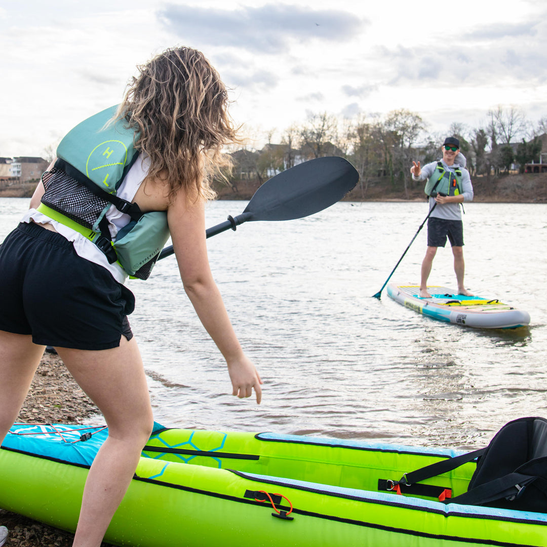 Woman approaches a man paddleboarding on a lake, holding a paddle near a kayak.