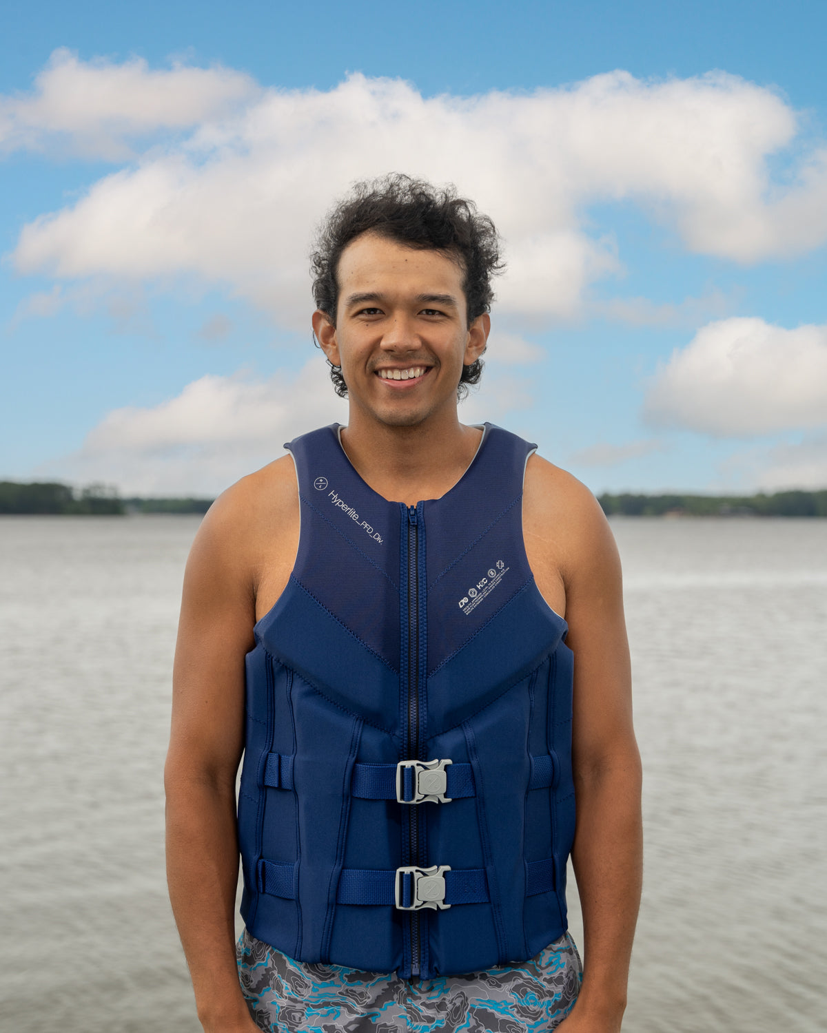 Person in blue life jacket smiles by the water with a cloudy sky in the background.