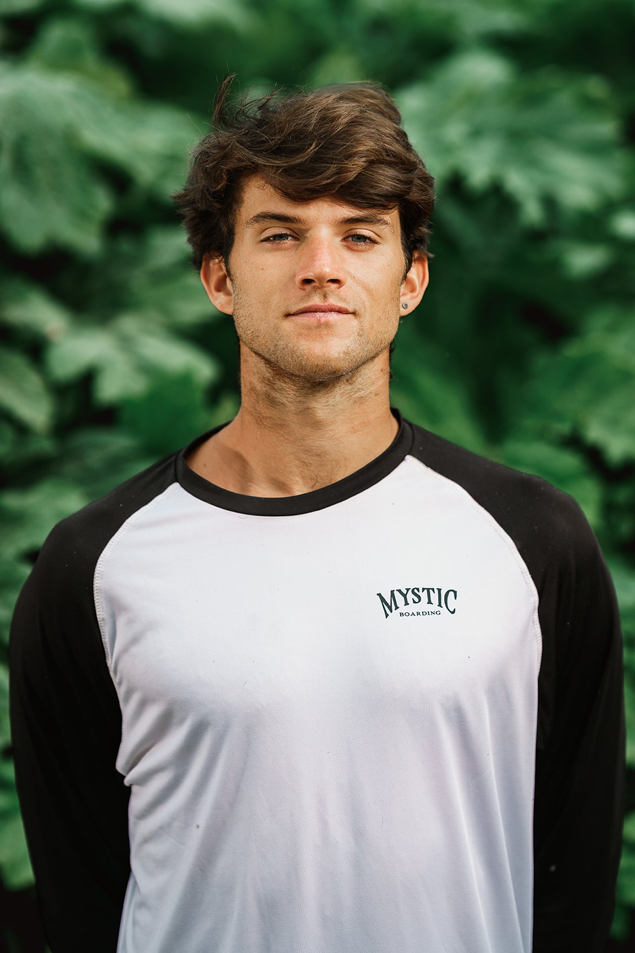 Person in a white and black shirt stands in front of lush green foliage.