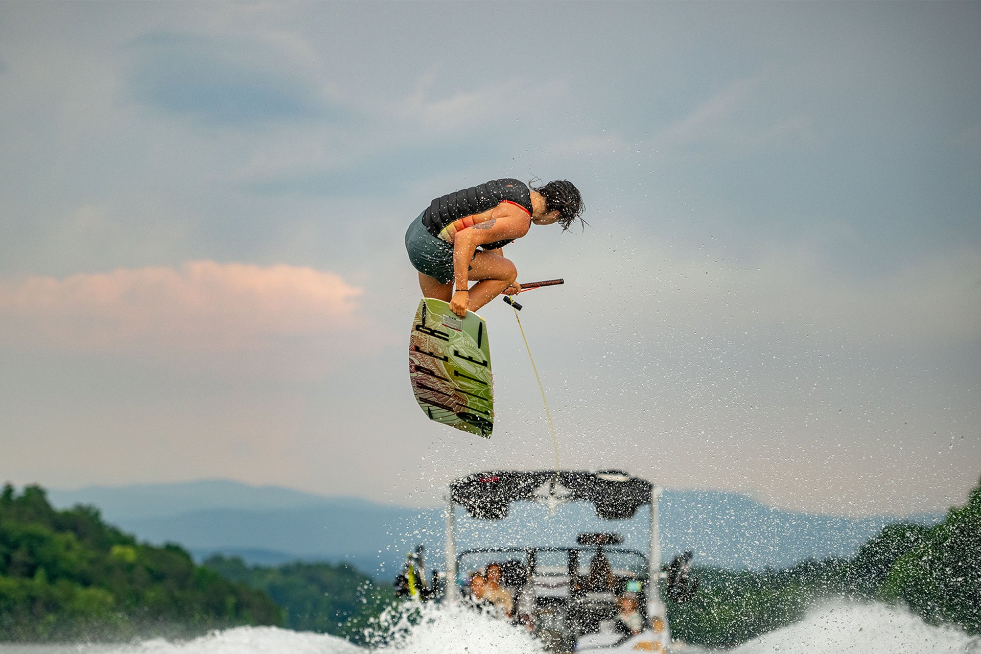 Person wakeboarding, performing a jump with a rope in hand, against a backdrop of trees and cloudy sky.