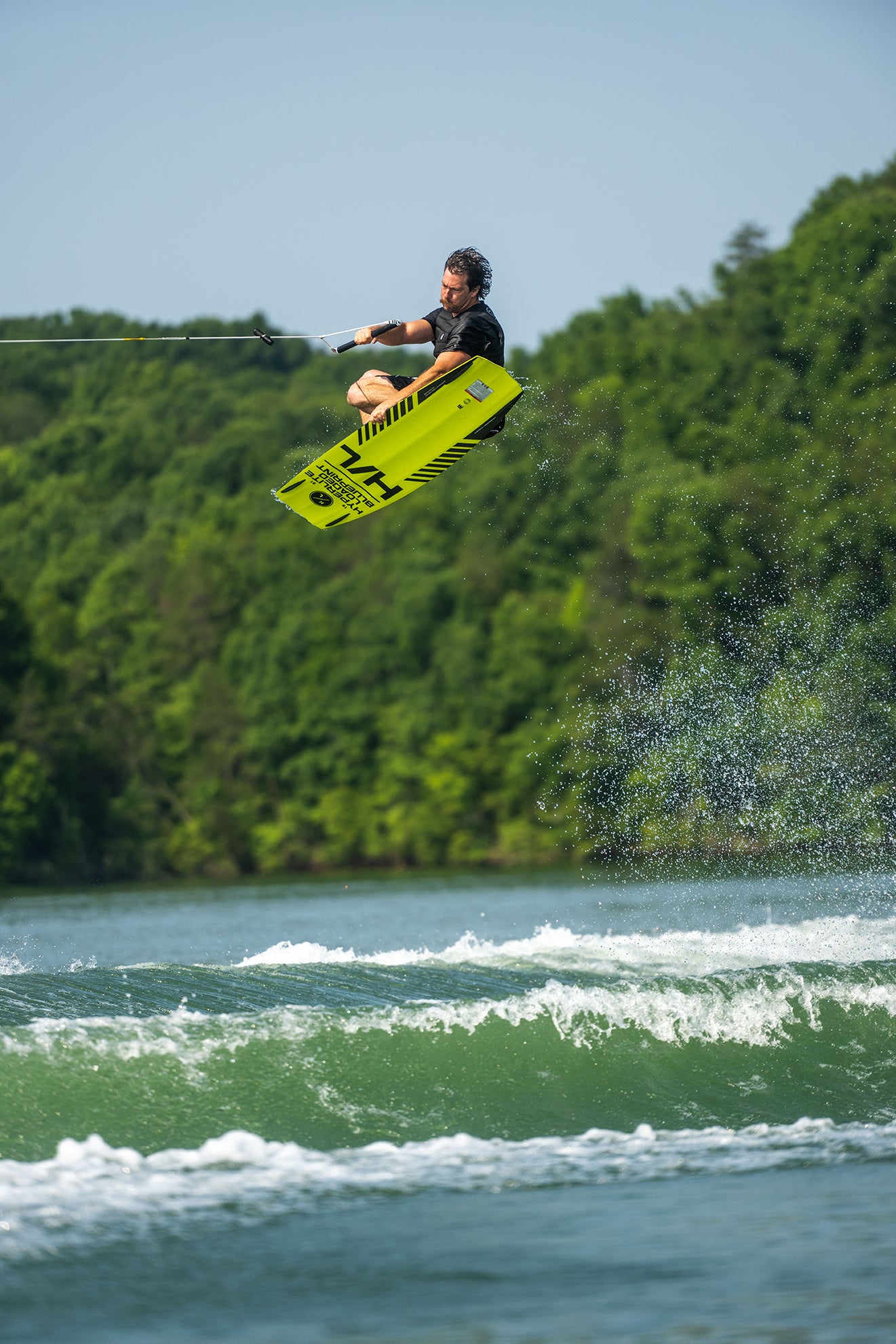 Person wakeboarding, mid-jump over water, surrounded by green trees.