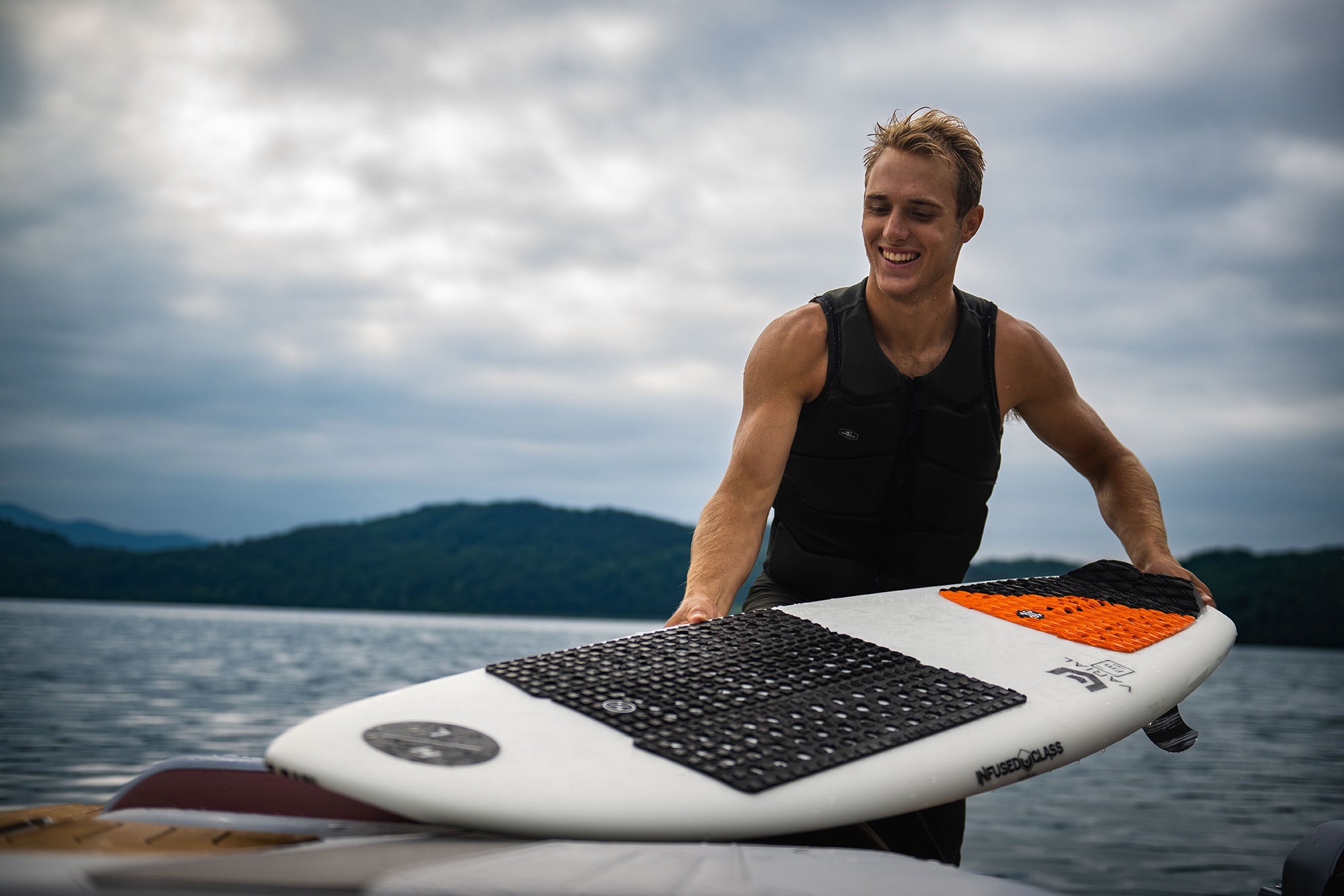 Person holding a surfboard on a lake with cloudy skies.