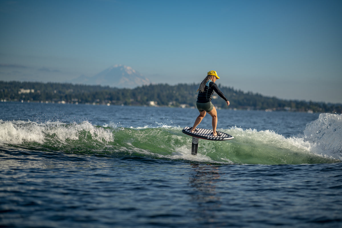 Person in a yellow hat foil surfing on a lake with a mountain in the background under a clear sky.