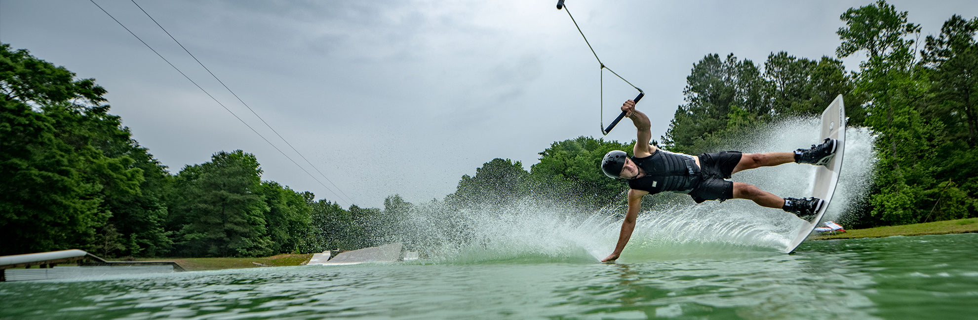 Person wakeboarding on a lake, performing a trick with one hand touching the water, surrounded by trees.