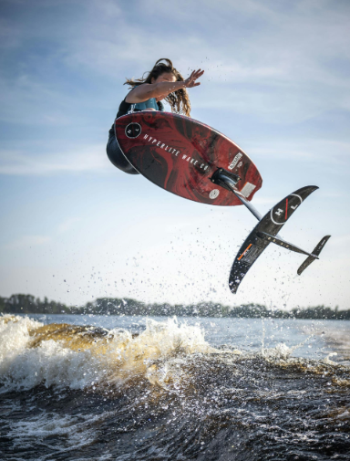 Person jumps on a hydrofoil surfboard over a wave under a clear sky.